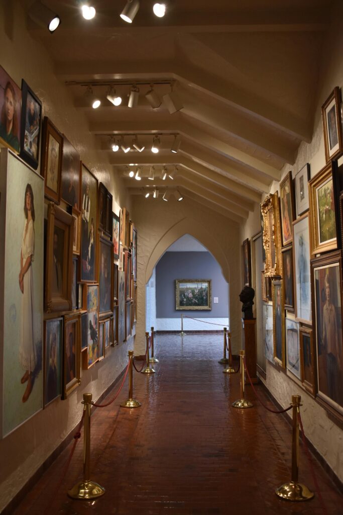 A narrow art gallery corridor showcasing diverse framed paintings under soft lighting.